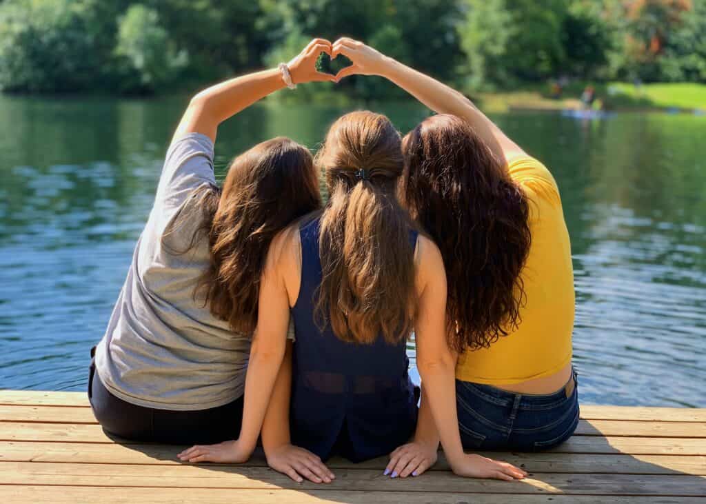 three teen girls making heart symbol with hands by lake