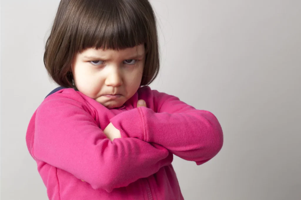 A young girl with a serious expression on her face, arms crossed in defiance, and wearing a bright pink sweater. This image represents a common challenge parents face when dealing with stubborn or defiant behavior and raises questions on how to spank effectively and responsibly.