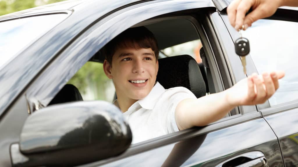Teen driver sits in driver seat as adult hands him the keys to the car
