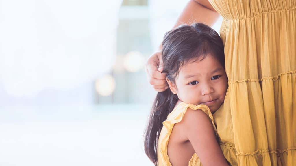 Close up of young, serious girl clinging to her mom's leg, with both wearing yellow summer dresses