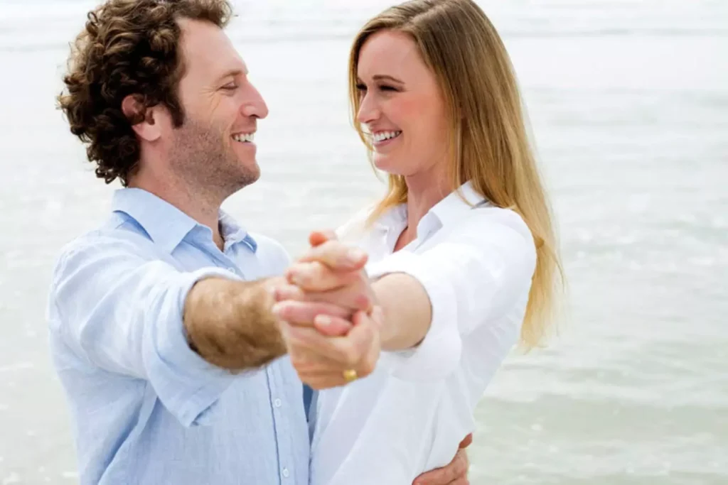 A smiling couple dancing near the ocean with the husband leading the dance, symbolizing how a husband can foster spiritual leadership in his home.