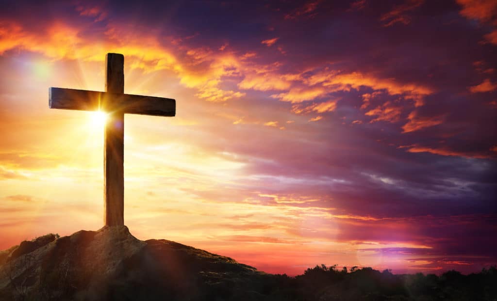 Photo of a cross on a hill, backlit by the sun.