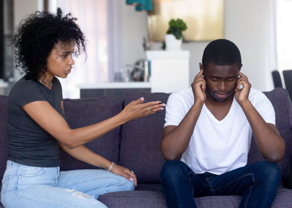 Controlling-black-wife-lecturing-her-black-husband-sitting-on-couch-with-hands-over-ears