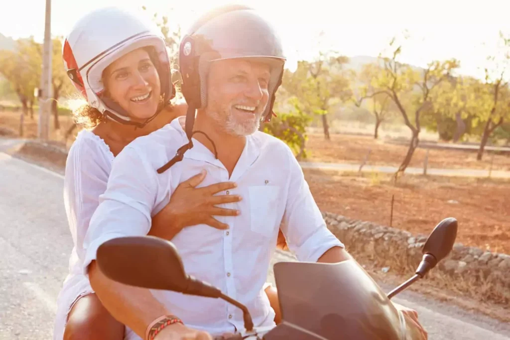 A joyful couple rides a scooter down a scenic road, sharing laughter with their spouse as they embrace the adventure together.