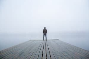 Young man standing alone on edge of footbridge and staring at lake. Mist over water. Foggy air. Early chilly morning in autumn. Beautiful freedom moment and peaceful atmosphere in nature. Back view.