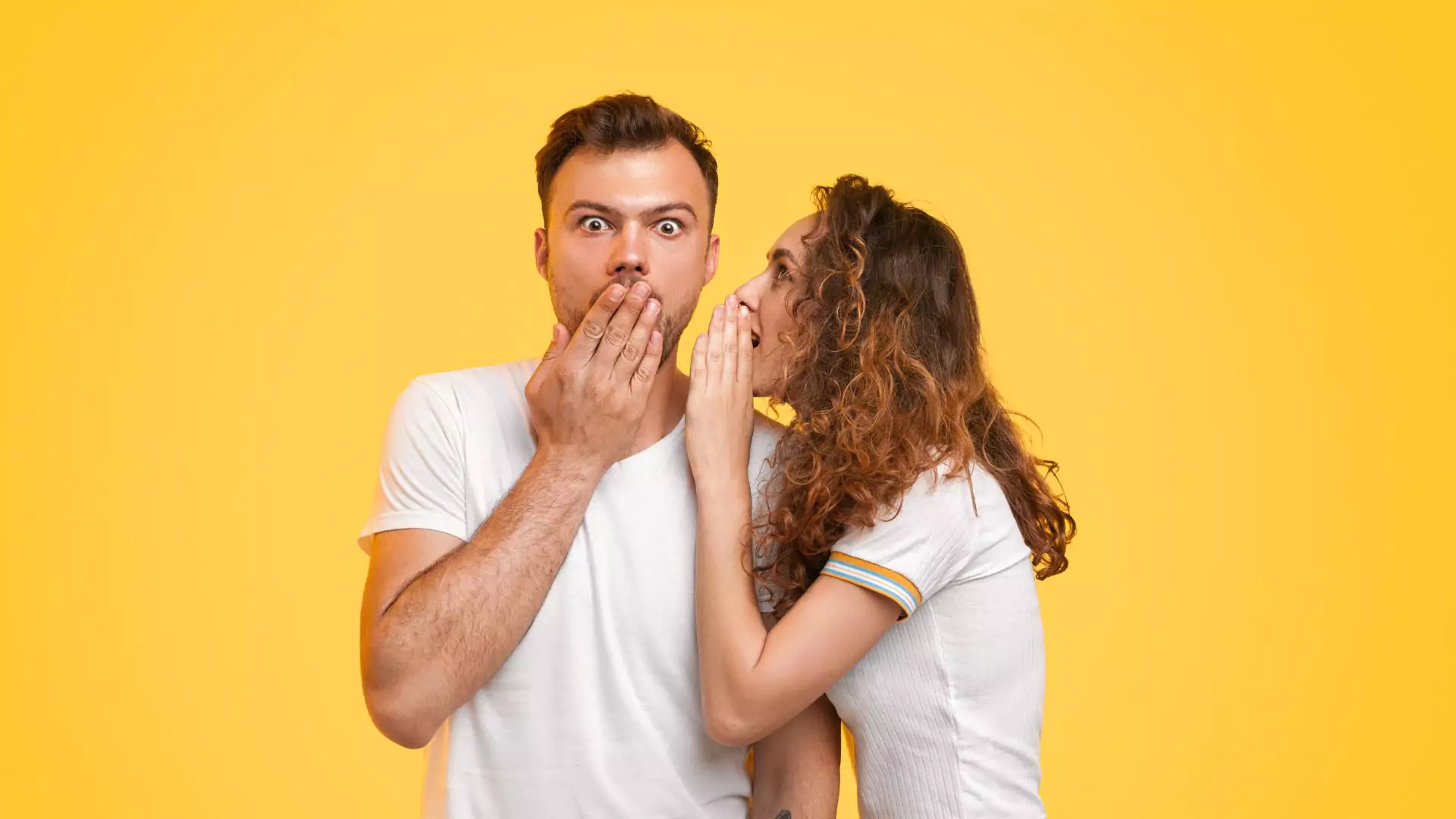 A young woman whispers into her fiancé’s ear while he looks surprised, representing the excitement and curiosity of asking important questions to ask your fiancé.