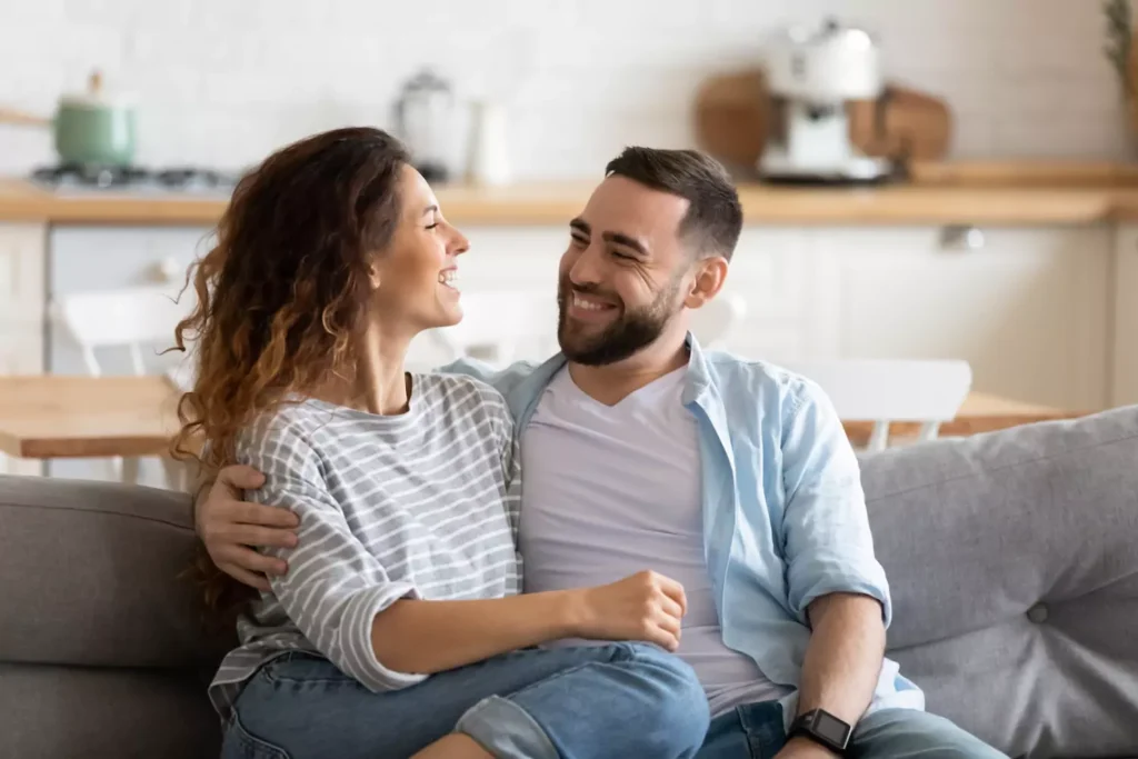 A young couple sit on the couch with their arms around each other. Validation in your marriage is key to conflict resolution.