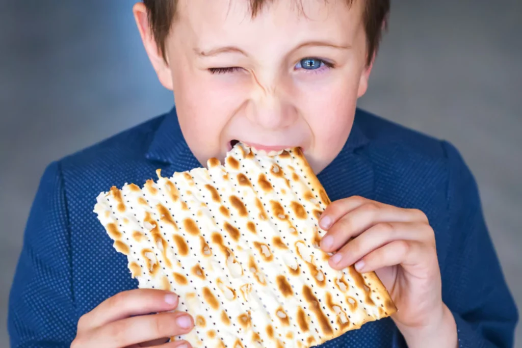 Young boy wearing a blue shirt, winking with one eye as he takes a big bite of a large piece of matzah bread.