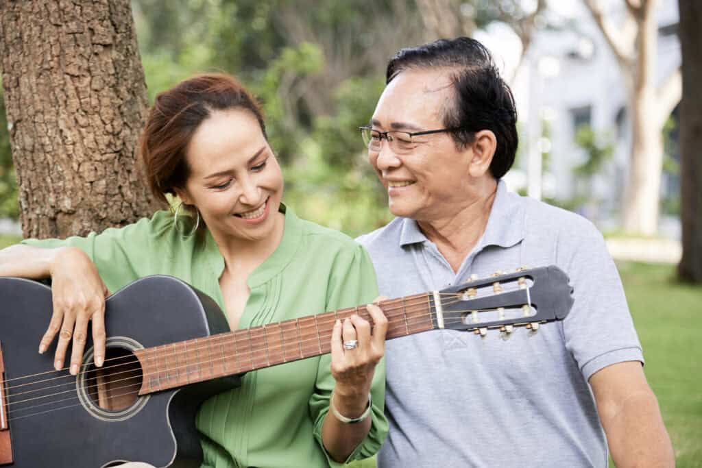 Proud affectionate senior man looking at his talented wife playing guitar and singing when they are resting in park.